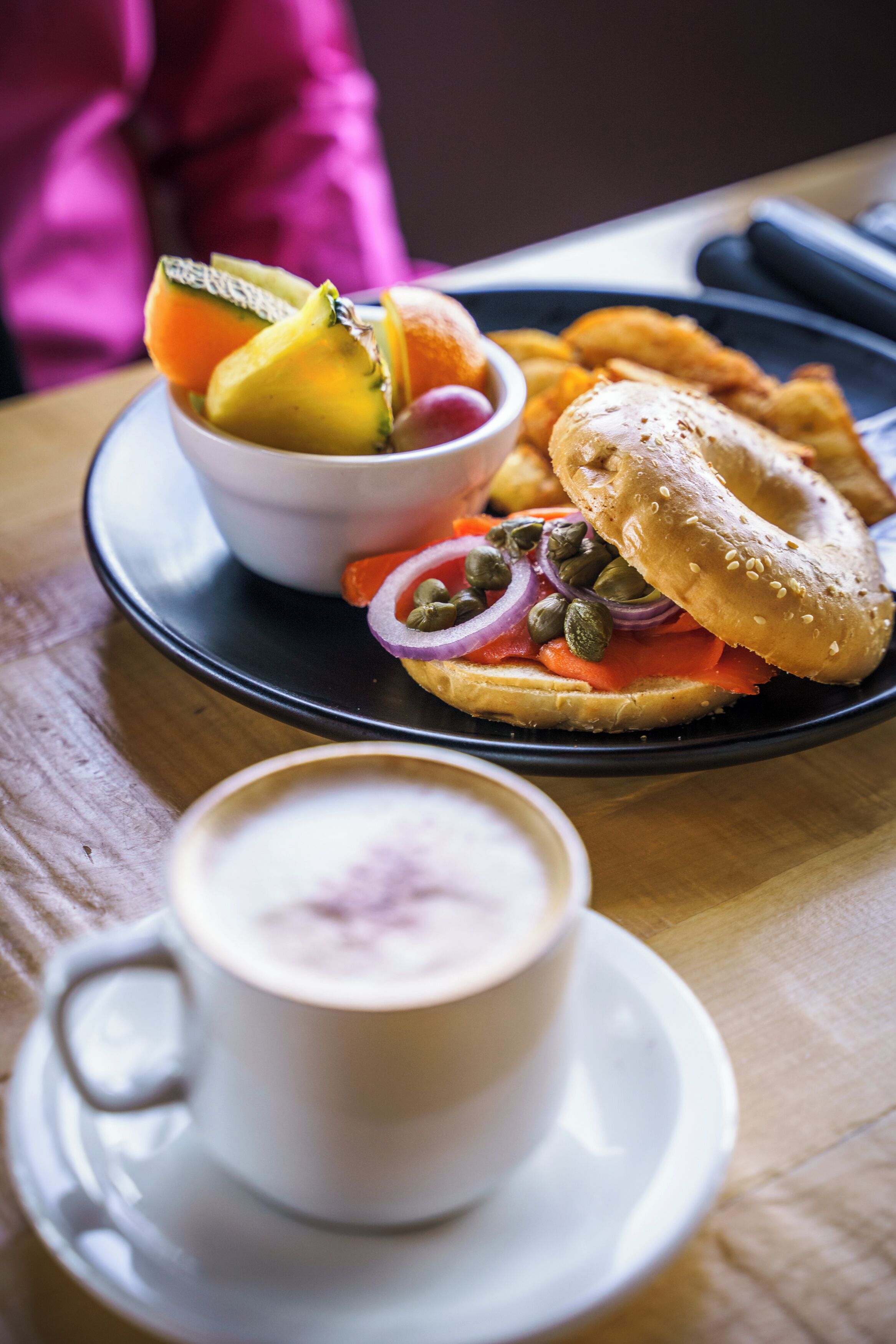 A breakfast plate with a bagel, smoked salmon, capers, red onion, and a bowl of fruit on a wooden table.