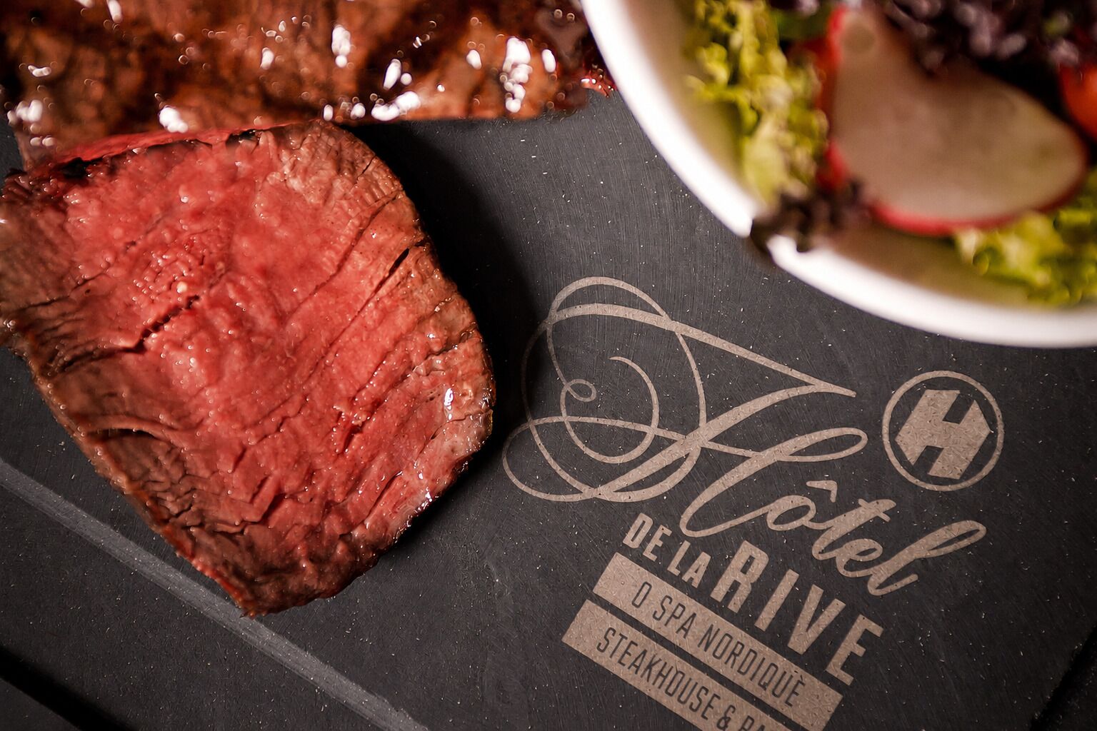 A steak on a black surface with a logo for 'Hotel de Marive SPA Nordique Steakhouse & Bar' and a bowl of salad.