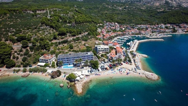 An aerial view of a coastal town with a beach, clear blue waters, and mountainous backdrop. - Sirena Hotel (Podgora)