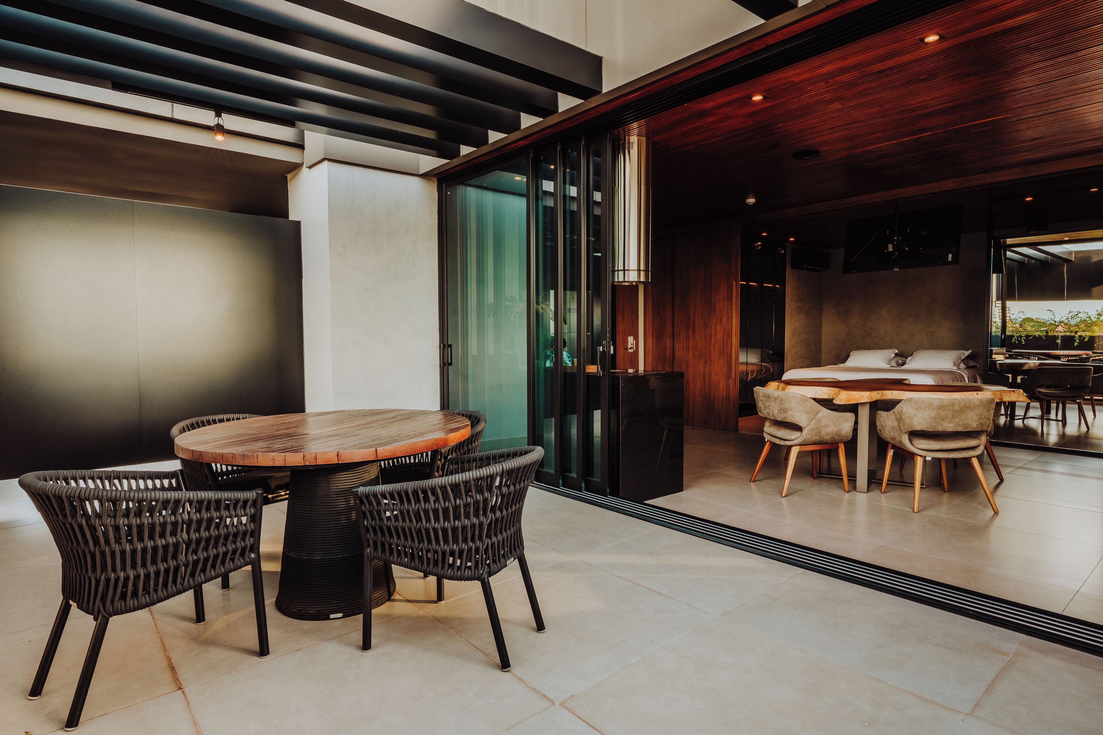 A modern dining area with a round wooden table and wicker chairs, a sofa set, and a large window offering a view of greenery.