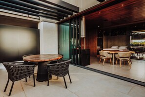 A modern dining area with a round wooden table and wicker chairs, a sofa set, and a large window offering a view of greenery. - Catuai Hotel (Cacoal)