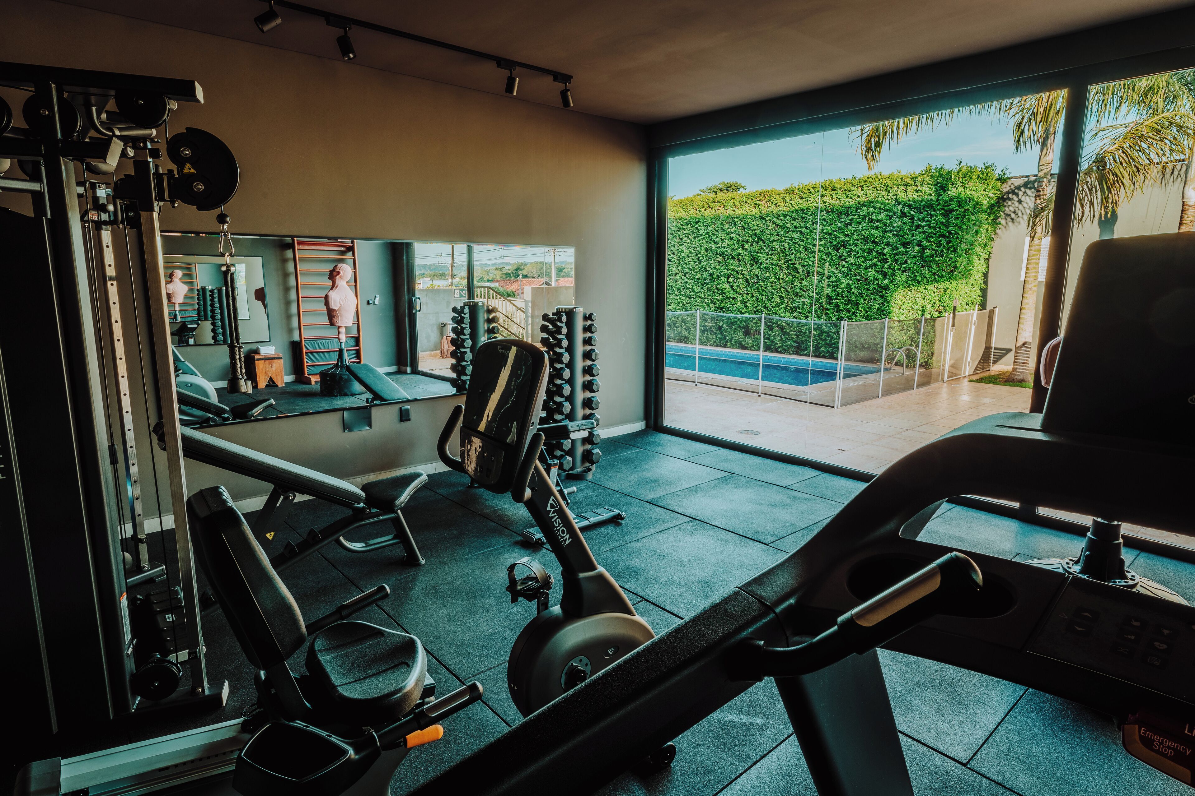 Une salle de sport moderne équipée de matériel d’exercice, d’un banc et offrant une vue sur une piscine et un espace verdoyant.