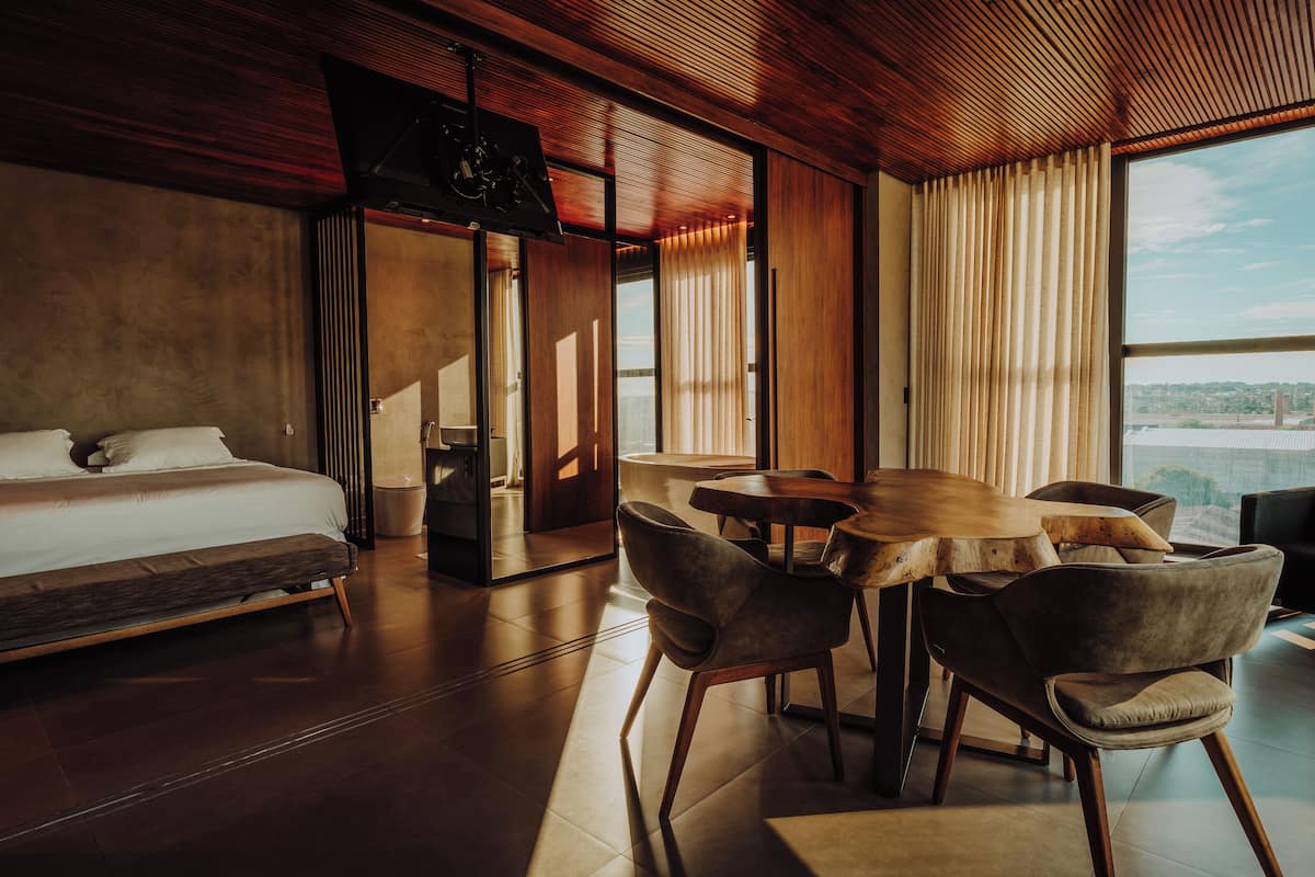 A modern hotel room with a large bed, a dining table with chairs, and a view of the city.