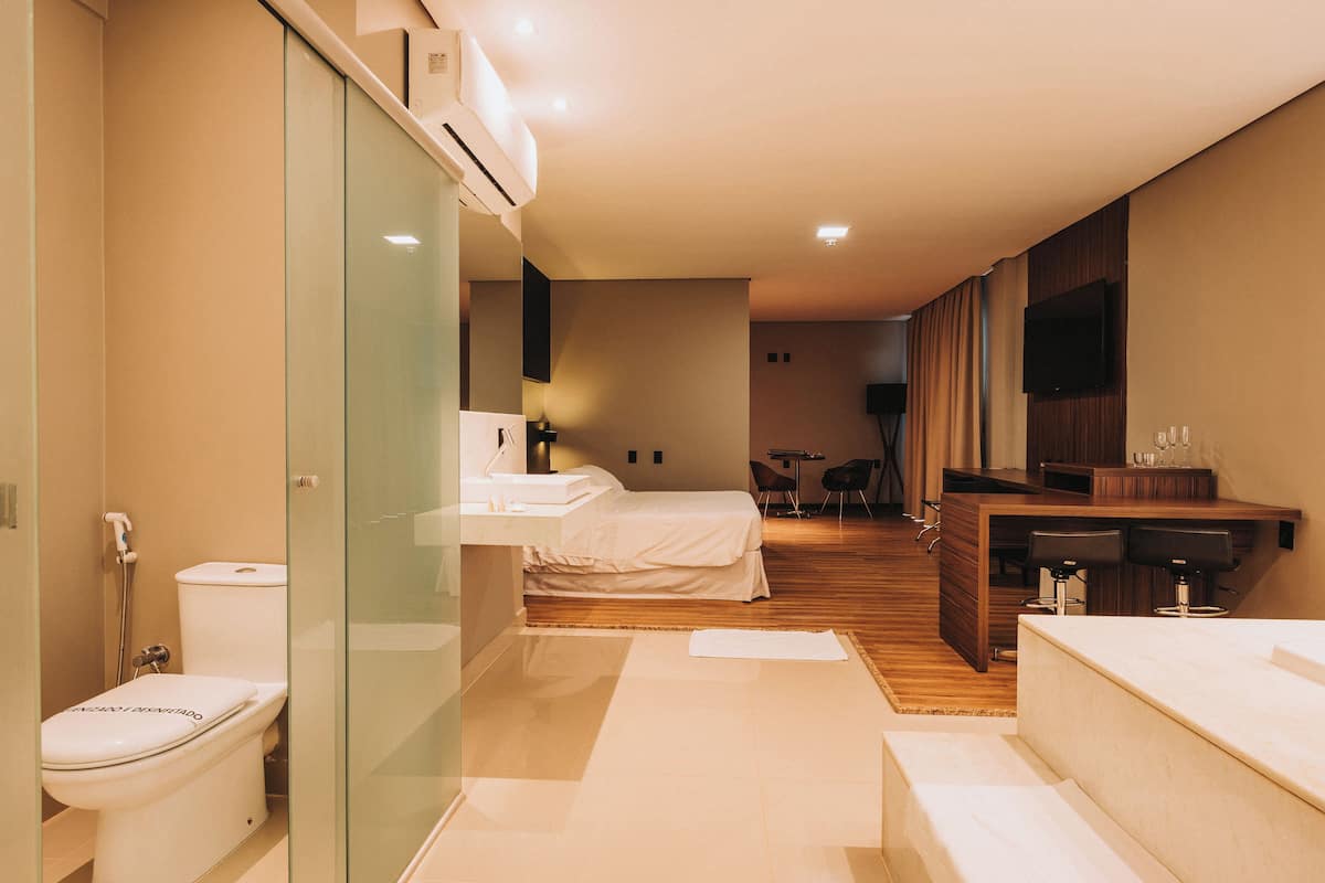 A modern hotel room with a glass-enclosed shower, a separate toilet, a bed, a small dining area, and a TV mounted on the wall.