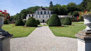 Un edificio grande de estilo clásico con una fachada simétrica, una entrada central y un jardín cuidado con setos podados y macizos de flores.