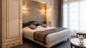 Una habitación de hotel con una cama grande, una mesita de noche, una mesa redonda con un florero y una ventana con cortinas.