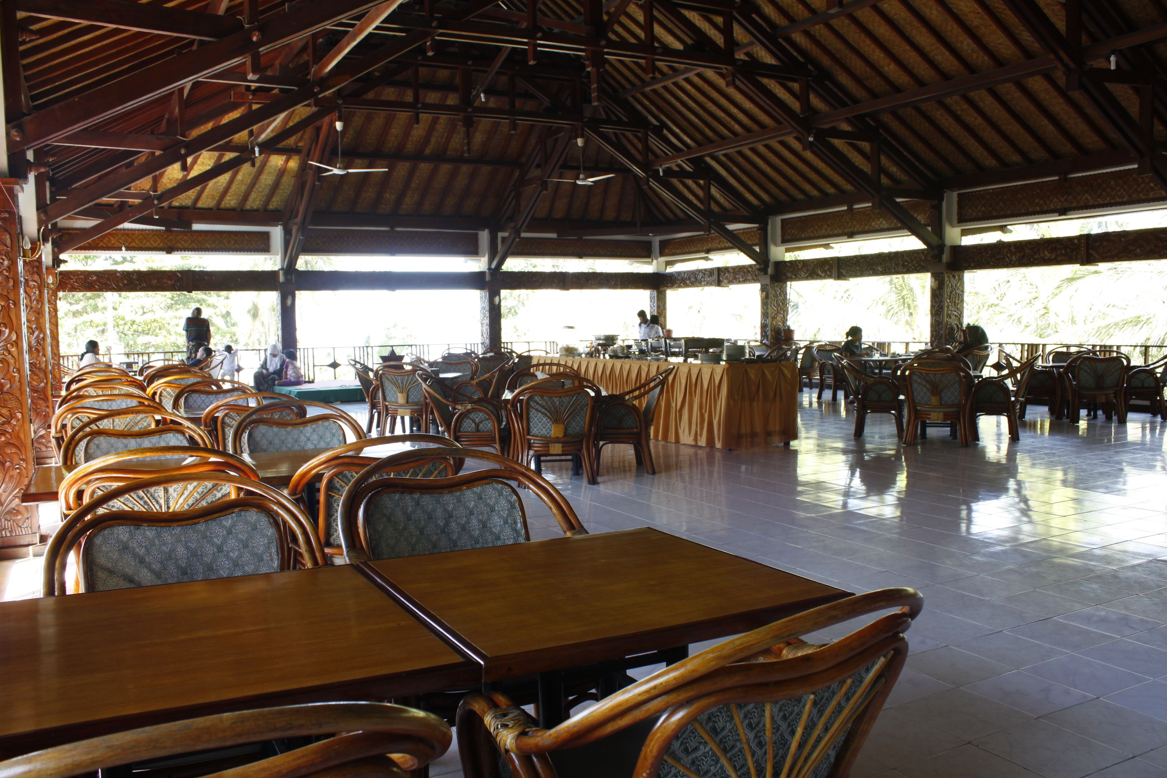 A spacious indoor dining area with wooden tables and chairs, a high ceiling, and large windows allowing natural light.