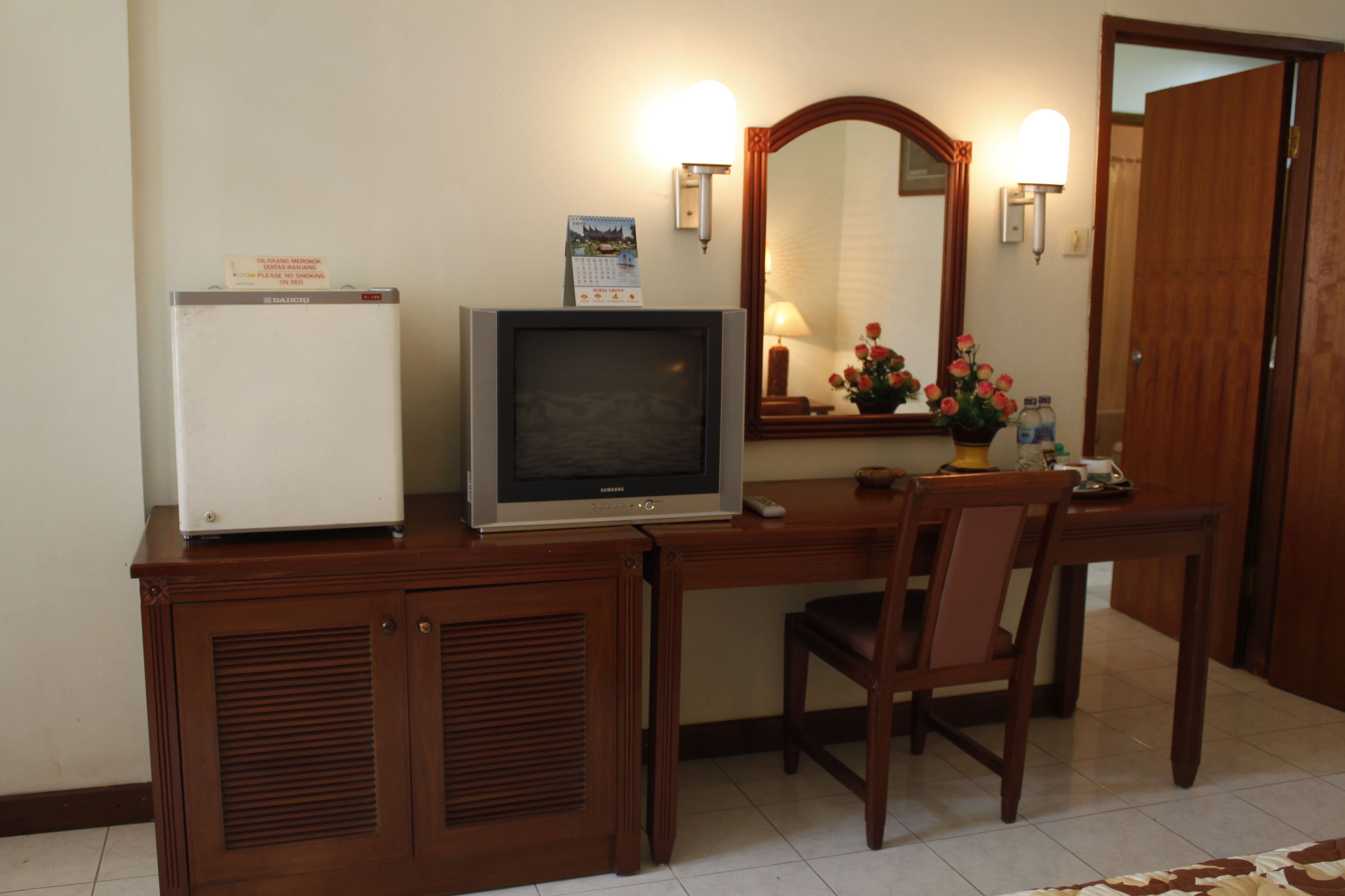 Una habitación de hotel pequeña con un escritorio de madera, televisor, refrigerador, espejo y puerta.