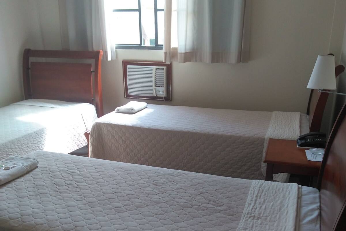 Una habitación de hotel con dos camas, una mesita de noche con lámpara, una ventana con cortinas y un aire acondicionado instalado en la pared.
