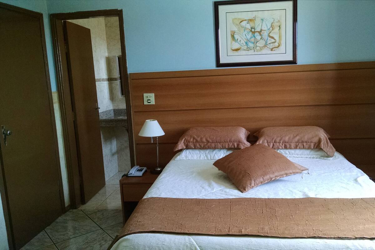 Habitación de hotel con cabecera de madera, cama con ropa blanca y almohadas marrones, lámpara de noche, teléfono y una obra de arte enmarcada en la pared.