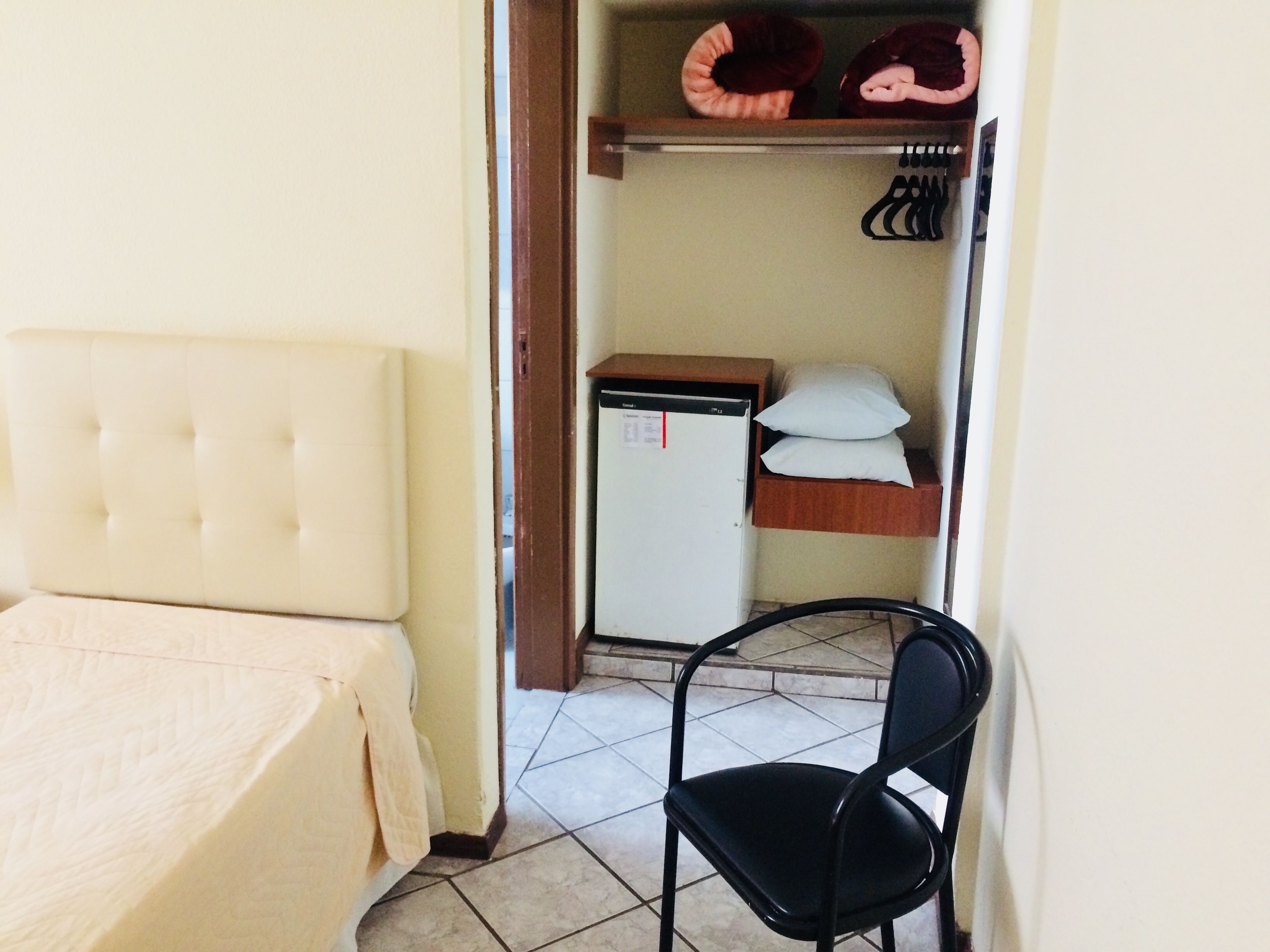 A bedroom with a bed, a small refrigerator, a chair, and a closet with hanging items.