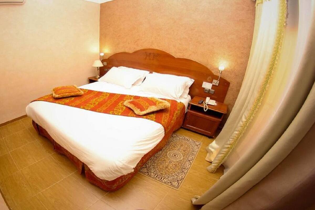 Quarto de hotel com uma cama grande, uma mesa de cabeceira, um abajur e um tapete estampado.