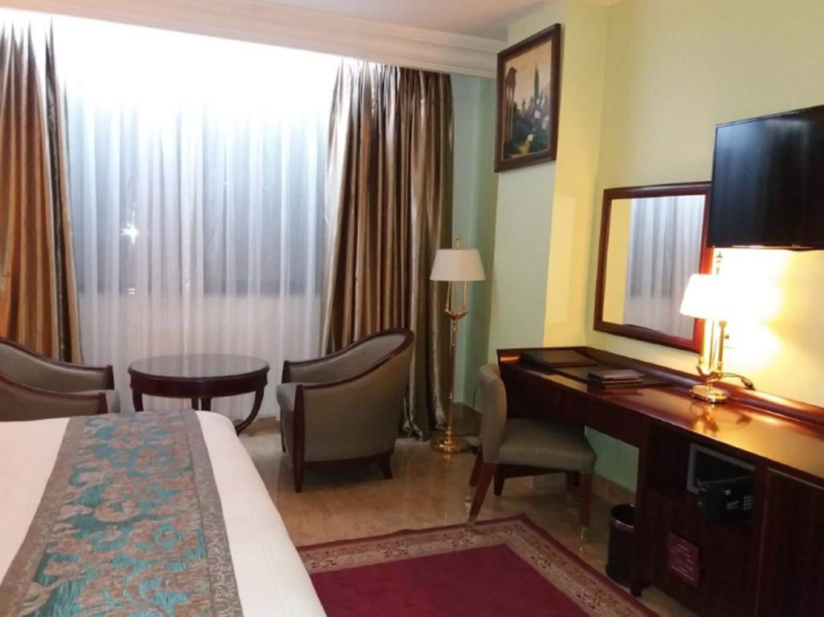 A hotel room with a bed, two chairs, a desk, a lamp, a television, and a painting on the wall.
