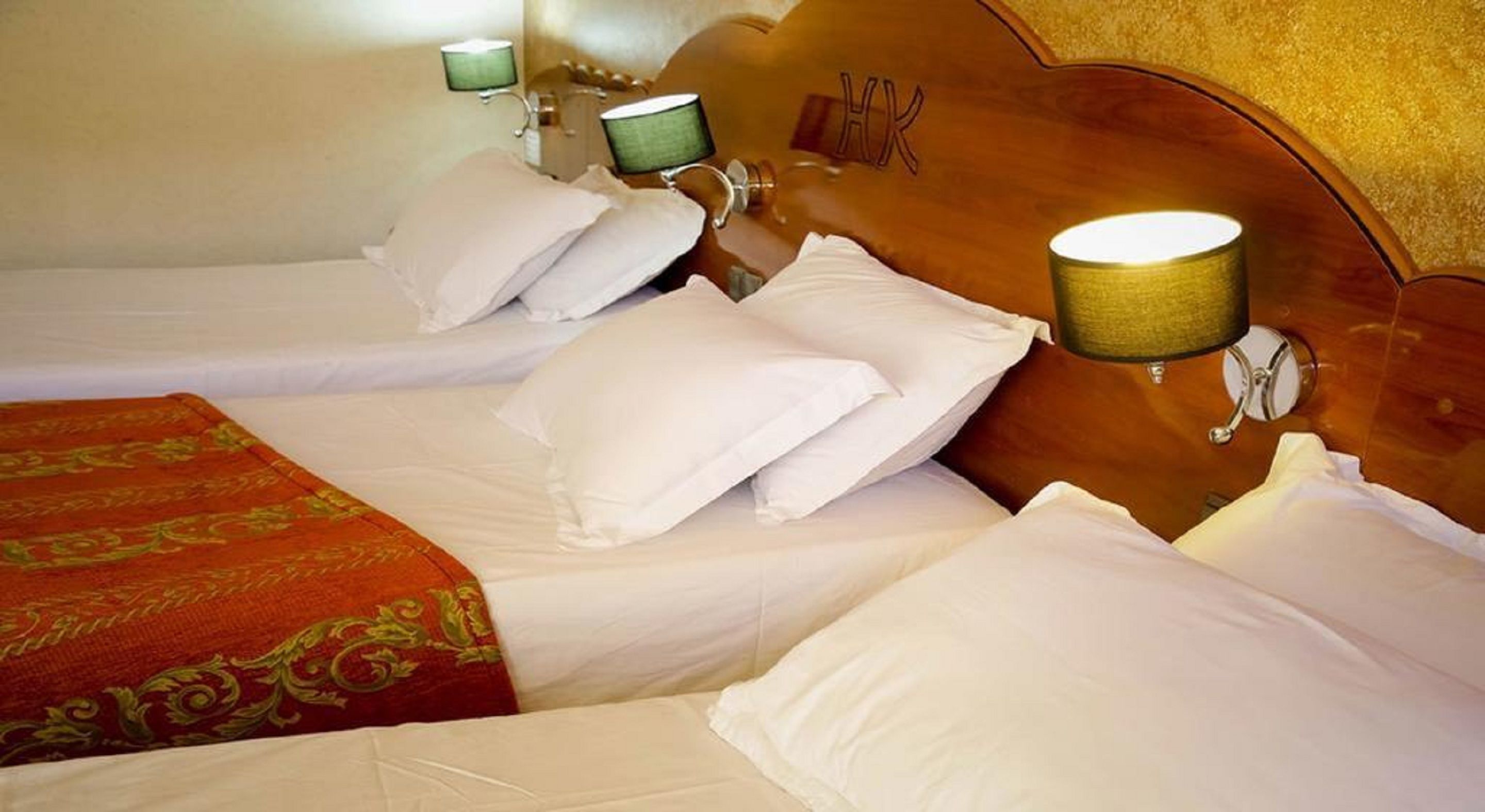 A hotel room with two beds, each with white linens and pillows, and a wooden headboard with a 'HOTEL' sign. There are two bedside lamps with green shades.