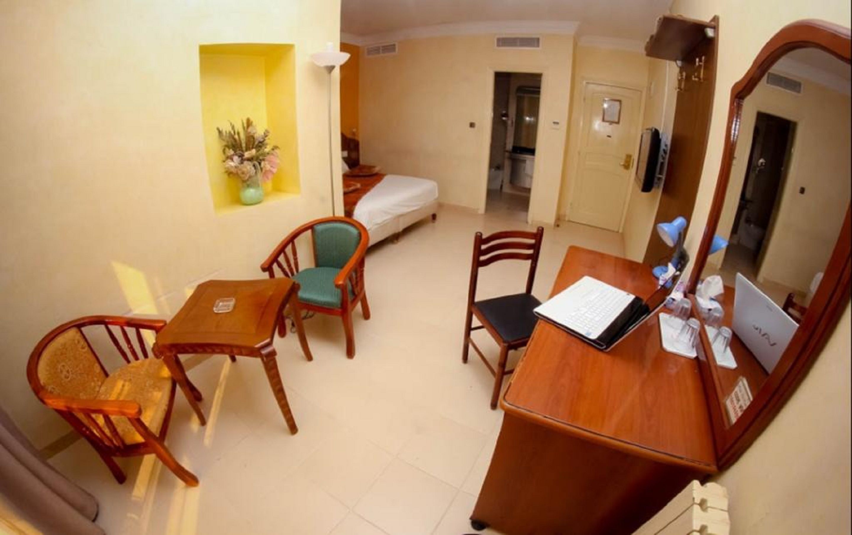 Habitación de hotel pequeña con cama, escritorio con una computadora portátil, dos sillas, una mesita, espejo y planta en maceta.