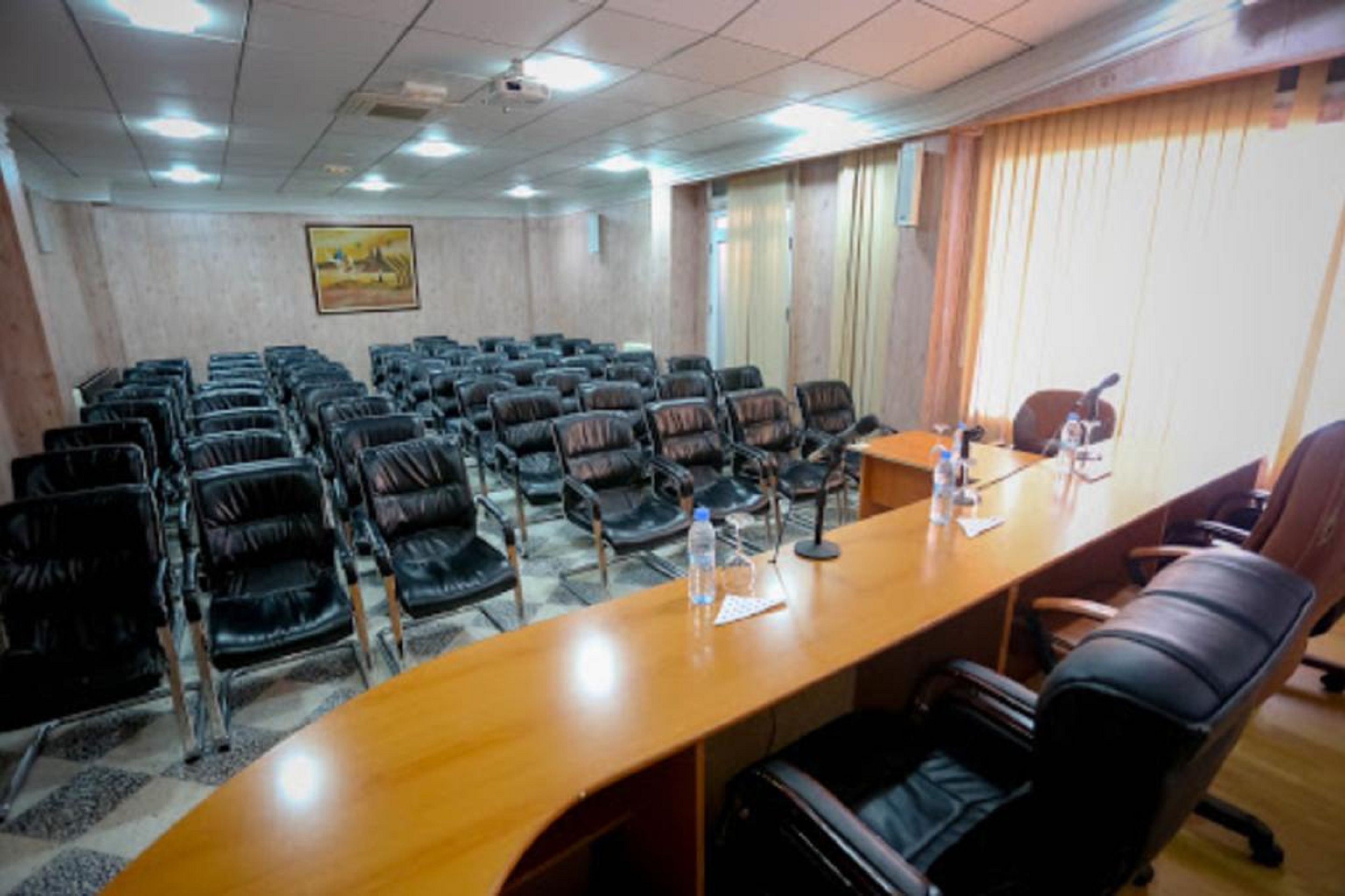 A conference room with a long wooden table, multiple black leather chairs, and a projector screen on the ceiling.
