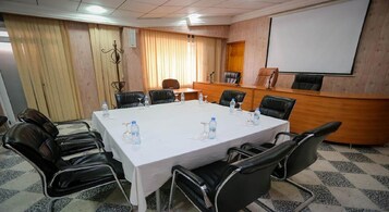 Un salón de conferencias con una gran mesa, sillas negras, botellas de agua, un atril y una pantalla de proyección.