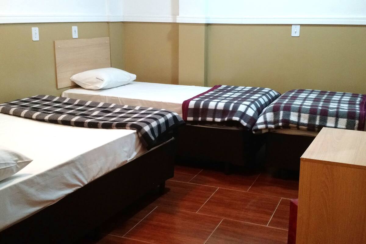 Een kamer met twee bedden, elk voorzien van een geruit plaid, een houten hoofdbord en een klein nachtkastje. De vloer is betegeld en de muren zijn beige.