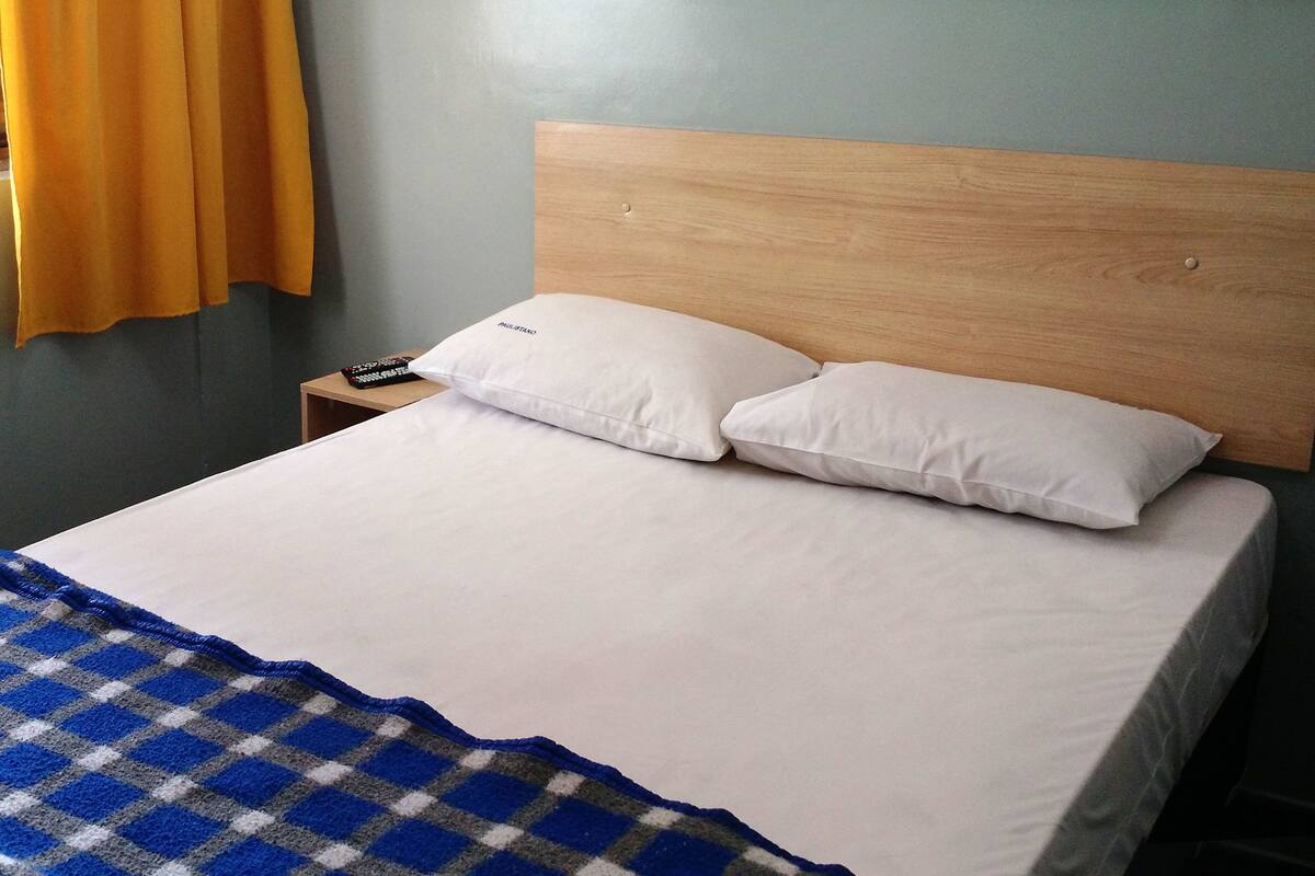 Een bed met witte beddengoed en twee kussens, een houten hoofdbord, een nachtkastje met een afstandsbediening, en een geruit plaid.