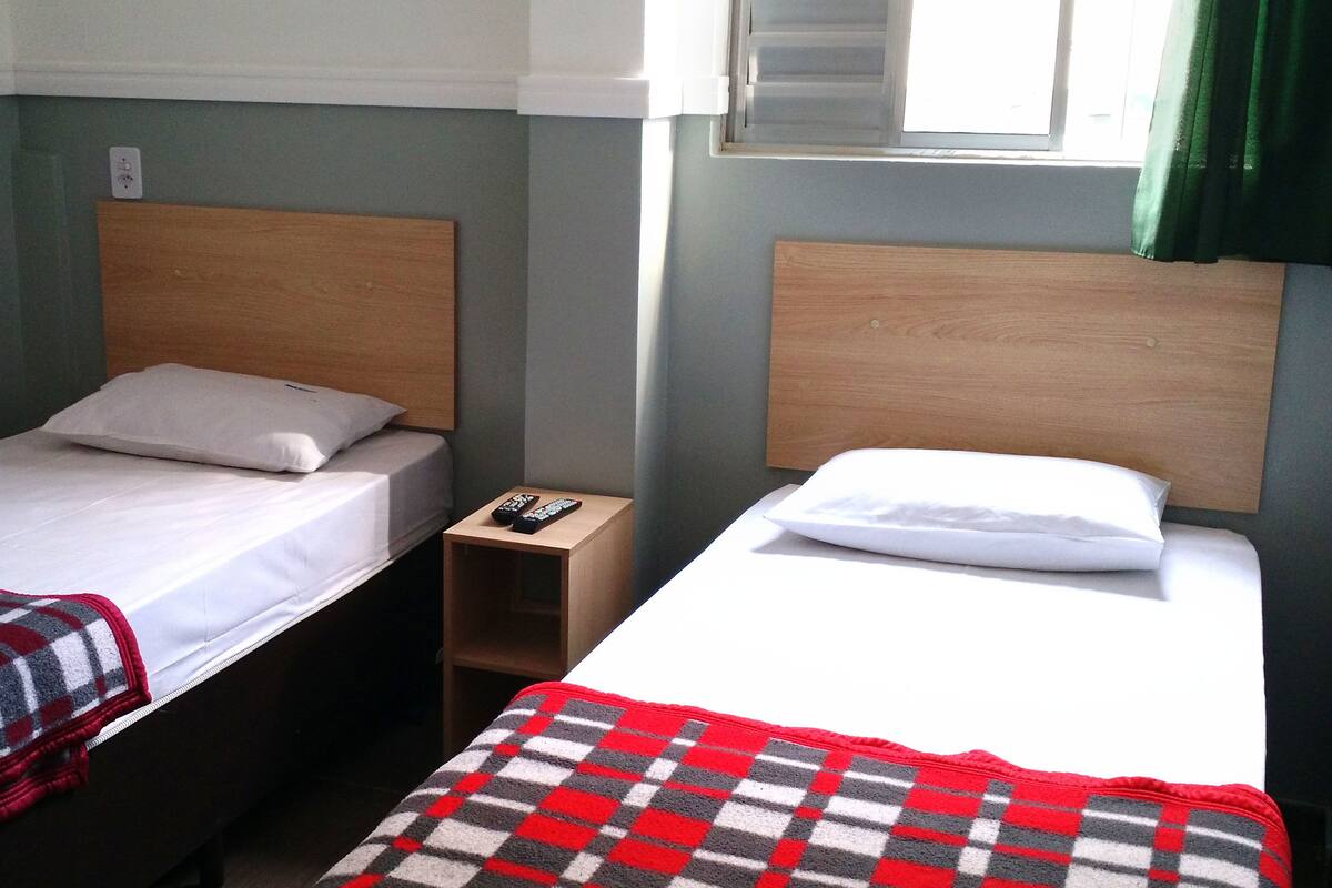 Twee enkele bedden met houten hoofdborden, een klein houten nachtkastje ertussen, en een rood-wit geruit kleed op de vloer.