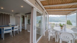 Un balcón amplio con techo de madera, mobiliario blanco y vistas a montañas y un lago.