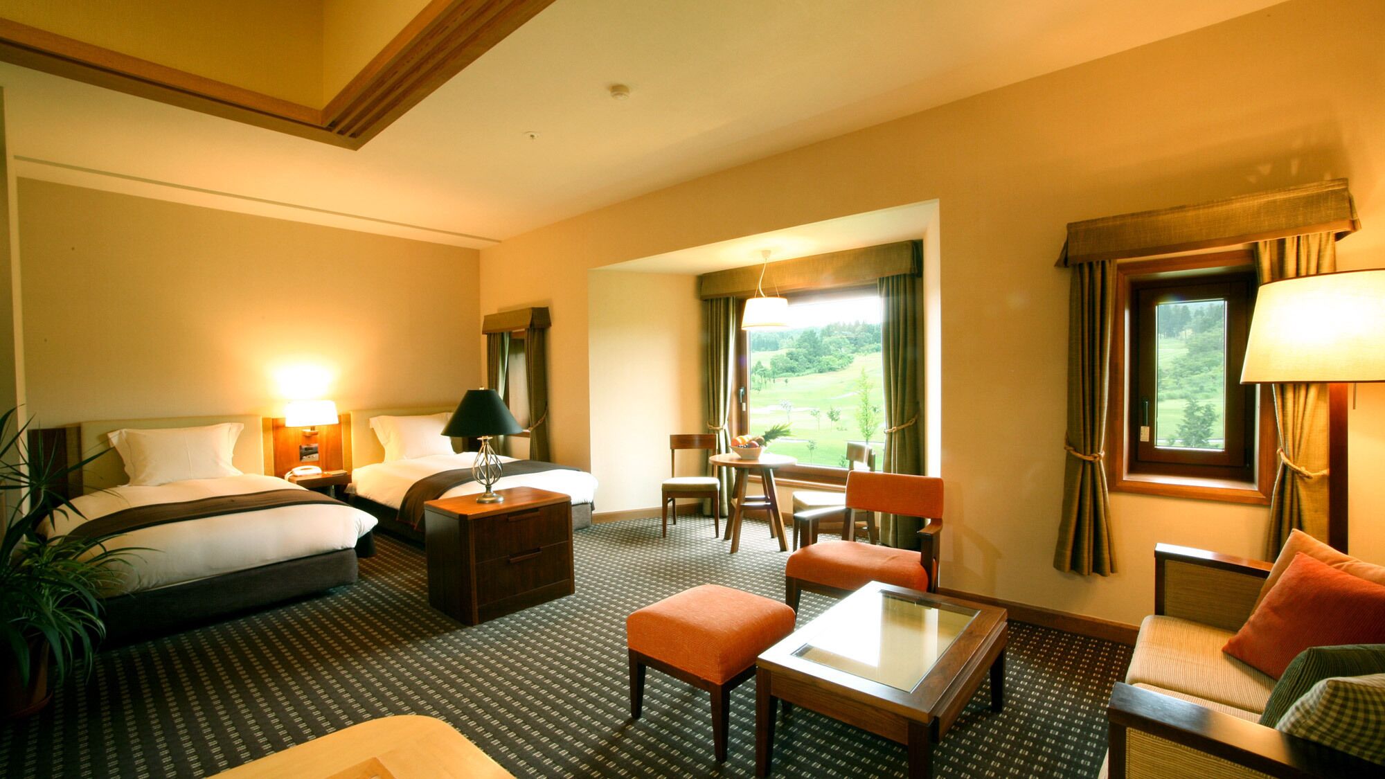 A hotel room with a bed, two bedside tables, a desk, a chair, an ottoman, a sofa, and a window with a view of greenery.