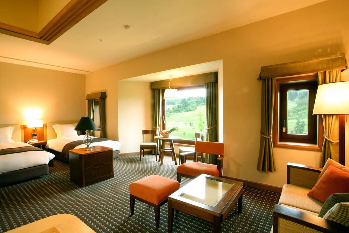 A hotel room with a bed, two bedside tables, a desk, a chair, an ottoman, a sofa, and a window with a view of greenery.