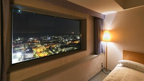 Habitación de hotel con un ventanal grande que da a un paisaje urbano nocturno, una lámpara de noche y una cama con cubrecama estampado.