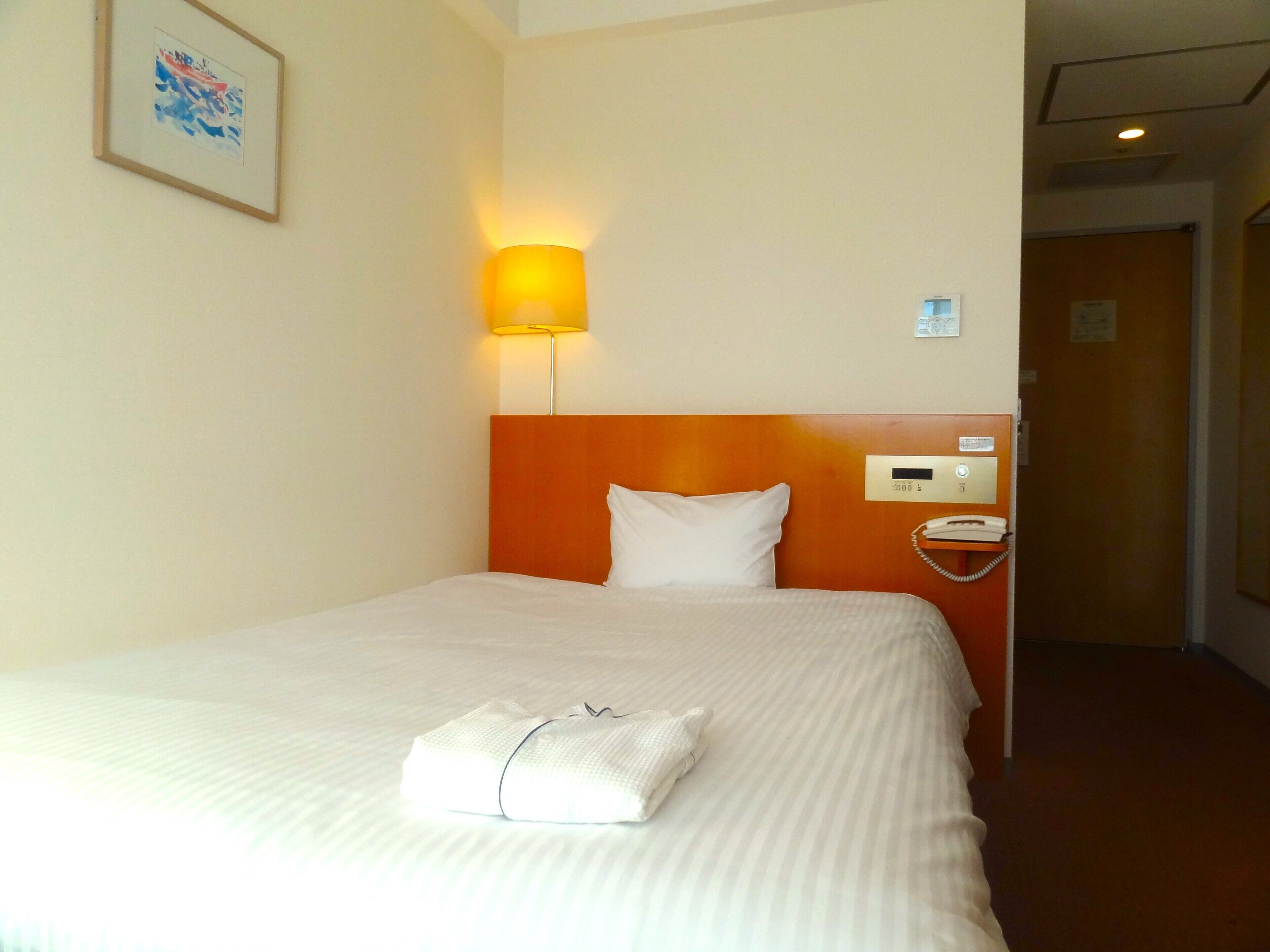 A hotel room with a neatly made bed, a bedside lamp, a telephone, and a framed picture on the wall.
