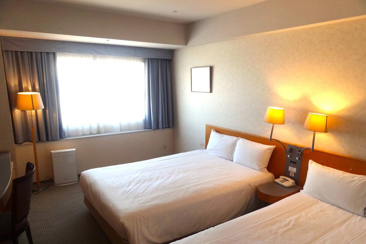 Habitación de hotel con dos camas, un escritorio, una silla, una lámpara y una ventana con cortinas.