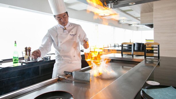 Un chef realizando una técnica culinaria con llamas sobre una plancha grande en una cocina moderna.