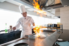 Ein Koch demonstriert eine Kochtechnik mit Flammen auf einer großen, flachen Kochplatte in einer modernen Küche.