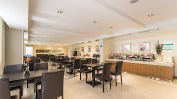 A modern hotel dining area with multiple tables and chairs, a central counter with food dispensers, and a clean, well-lit interior.