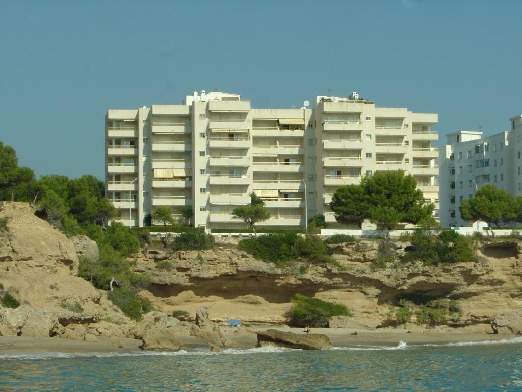 Un edificio de varios pisos con balcones, ubicado en una costa rocosa con una playa al frente.
