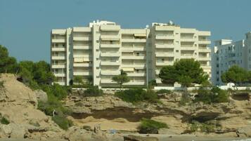 Un edificio de varios pisos con balcones, ubicado en una costa rocosa con una playa al frente.