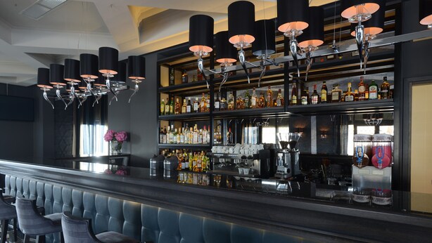 Uma área de bar com uma fileira de banquetas acolchoadas azuis, diversos frascos de bebidas alcoólicas nas prateleiras e uma máquina de café.