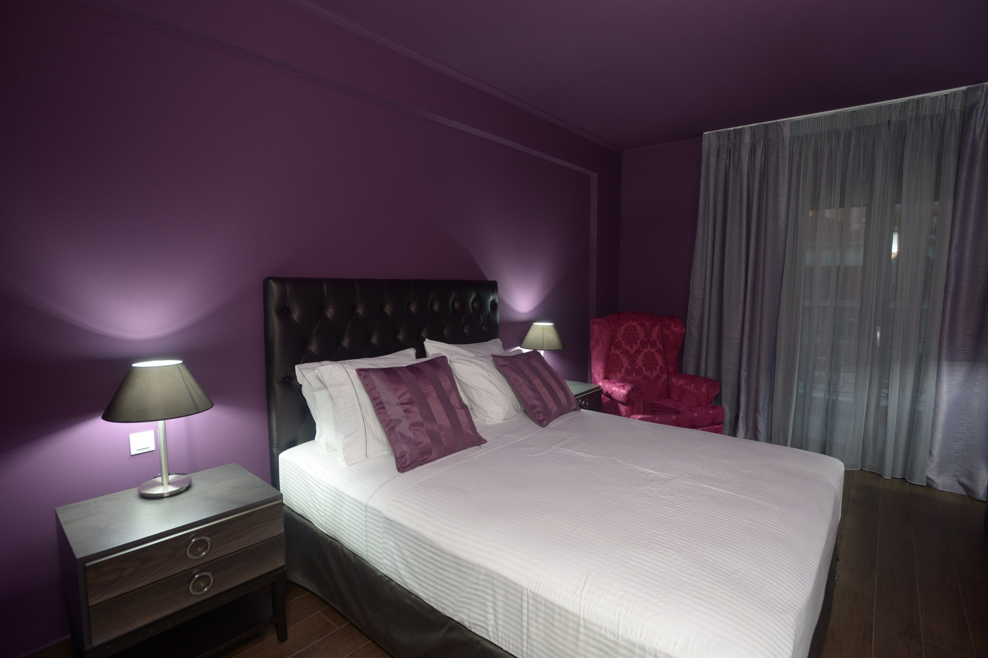 A neatly made bed with a headboard, two bedside lamps, a nightstand with drawers, and a red chair.