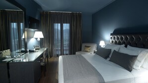 Quarto de hotel com uma cama grande, uma escrivaninha com luminária e vista para a cidade pelas janelas.