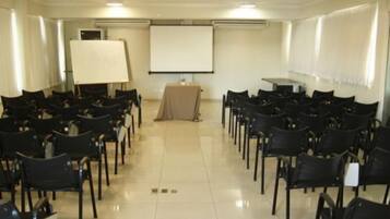 Una sala de conferències amb files de cadires negres, una pantalla de projecció blanca i una taula amb funda beix.