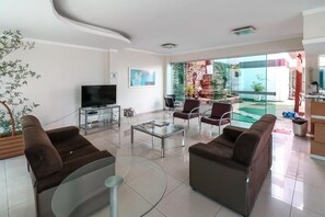 A modern living room with a flat-screen TV, two sofas, a glass coffee table, and a large sliding glass door leading to a balcony with a view of a garden and a building. - Turis Hotel (Campo Grande)
