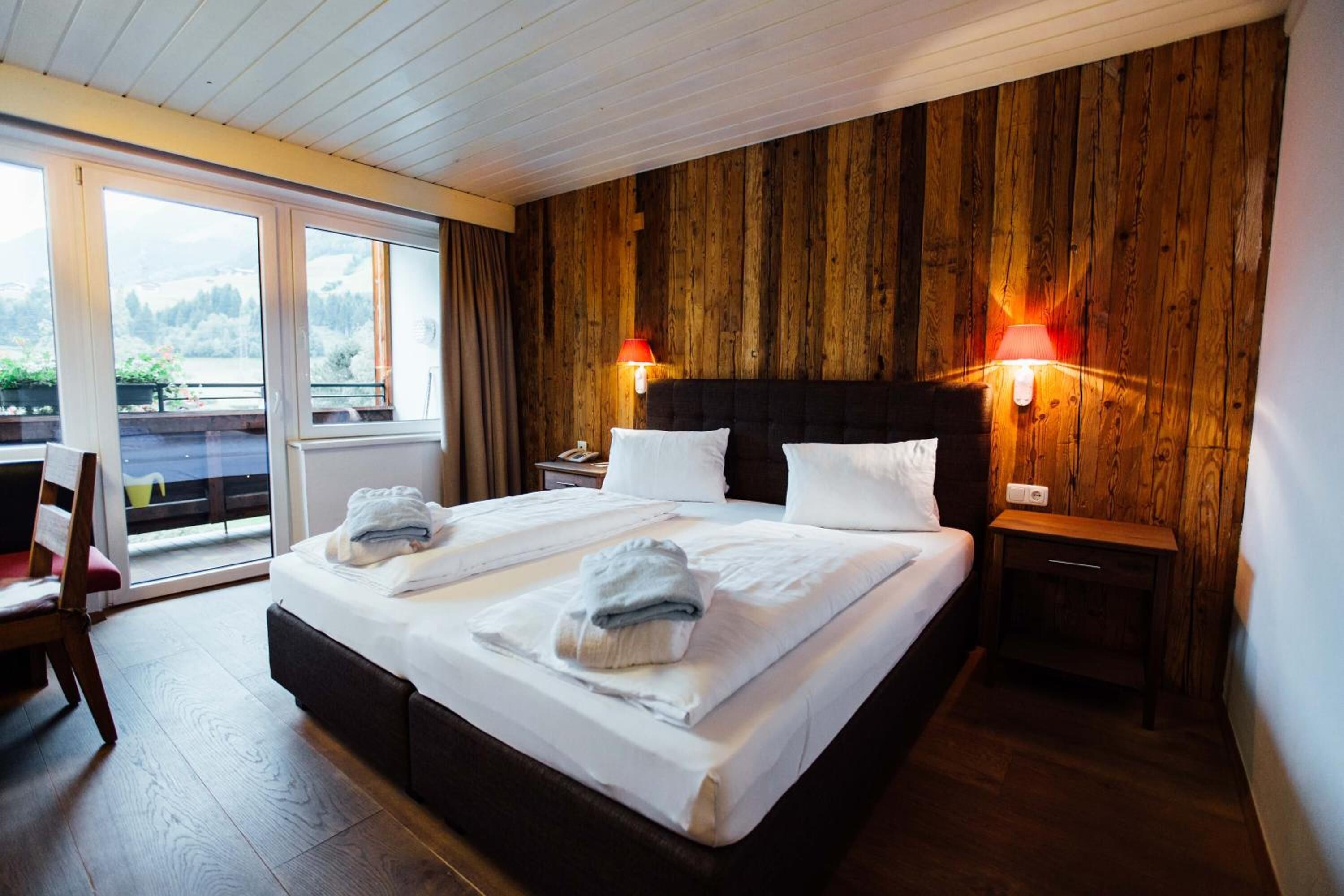 A bedroom with a wooden wall, a large bed with white linens, two bedside lamps, and a view through a sliding door.