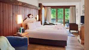 Een goed verlichte slaapkamer met een groot bed, houten lambrisering, een bureau met stoel, en een balkon met uitzicht op groen.
