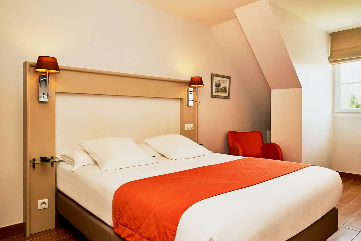 Un lit bien fait, recouvert d’une couvre-lit blanche et orange, deux lampes fixées au mur, un tableau encadré et une chaise rouge placée dans un coin.