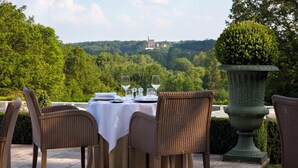 Un área de comedor al aire libre con sillas de mimbre y una mesa lista para dos, con vistas a un paisaje exuberante y un castillo distante.