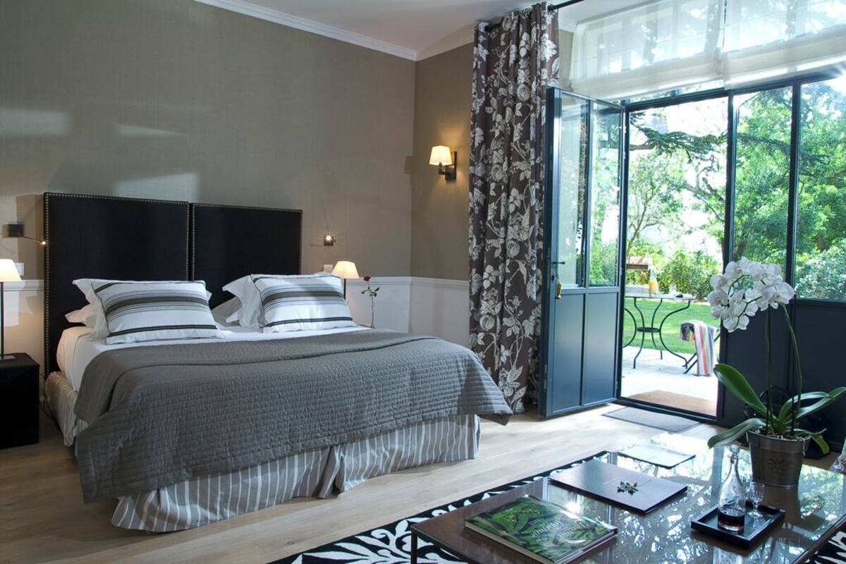 A bedroom with a bed, bedside lamps, a glass table with a tray, and a view of greenery outside.