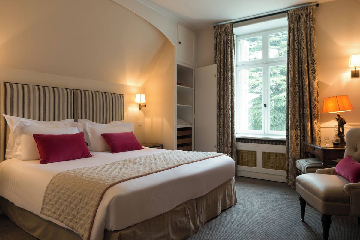 Una habitación de hotel con cama, una silla, una lámpara, una ventana y una estantería.