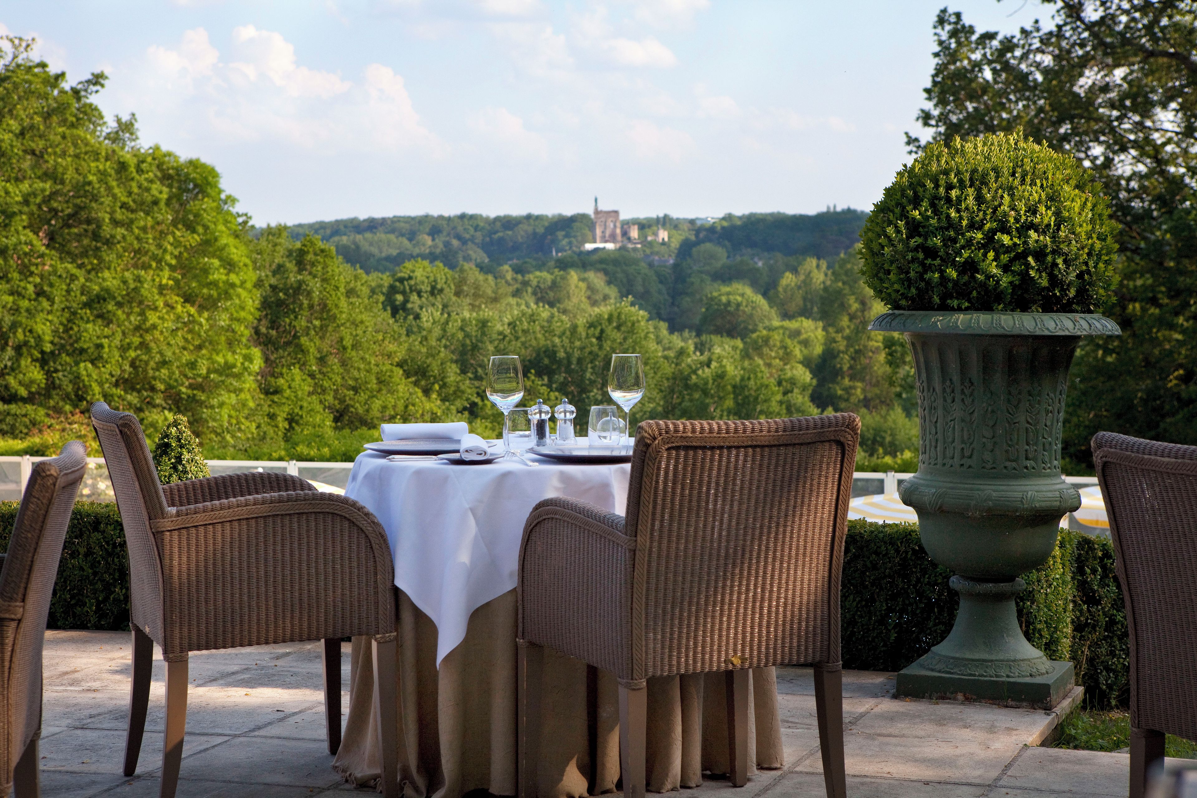 An outdoor dining area with wicker chairs and a table set for two, overlooking a lush landscape and distant castle.