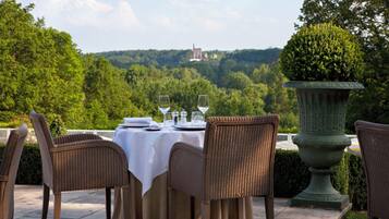 An outdoor dining area with wicker chairs and a table set for two, overlooking a lush landscape and distant castle.