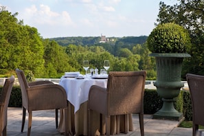 Un área de comedor al aire libre con sillas de mimbre y una mesa lista para dos, con vistas a un paisaje exuberante y un castillo distante.