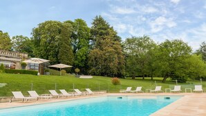 A swimming pool with lounge chairs, a covered seating area, and a building with a garden in the background. - Domaine de la Tortinière (Veigne)